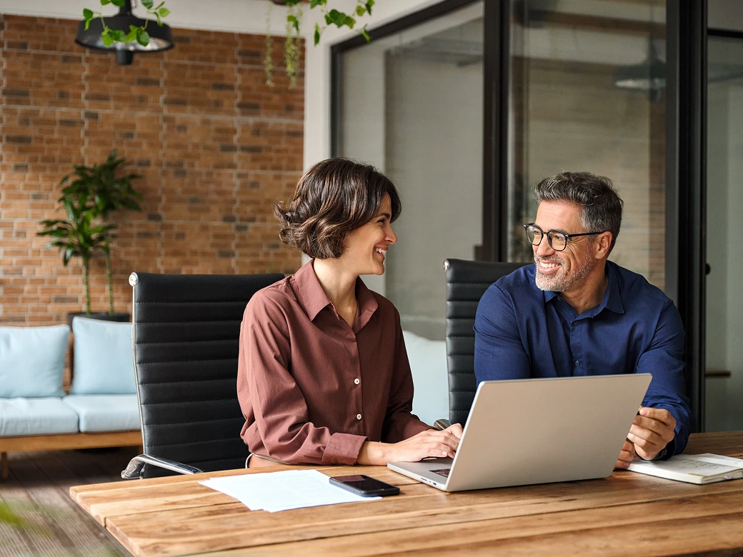 Two business professionals sit at a desk with a laptop, discussing work in a modern office setting.