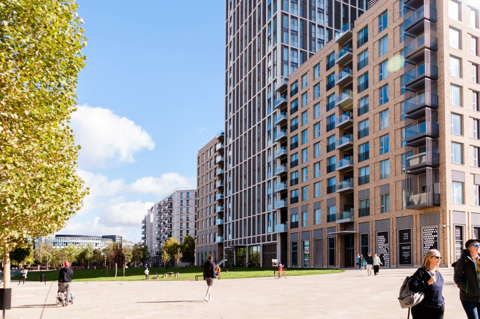 A modern urban scene featuring tall apartment buildings with large windows and numerous balconies. People are walking and socializing in a spacious plaza bordered by green lawns and trees, under a bright blue sky with a few scattered clouds. The contemporary architecture and outdoor space convey a vibrant, residential community atmosphere.