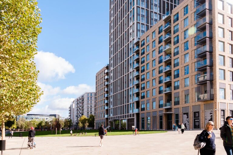 A modern urban scene featuring tall apartment buildings with large windows and numerous balconies. People are walking and socializing in a spacious plaza bordered by green lawns and trees, under a bright blue sky with a few scattered clouds. The contemporary architecture and outdoor space convey a vibrant, residential community atmosphere.