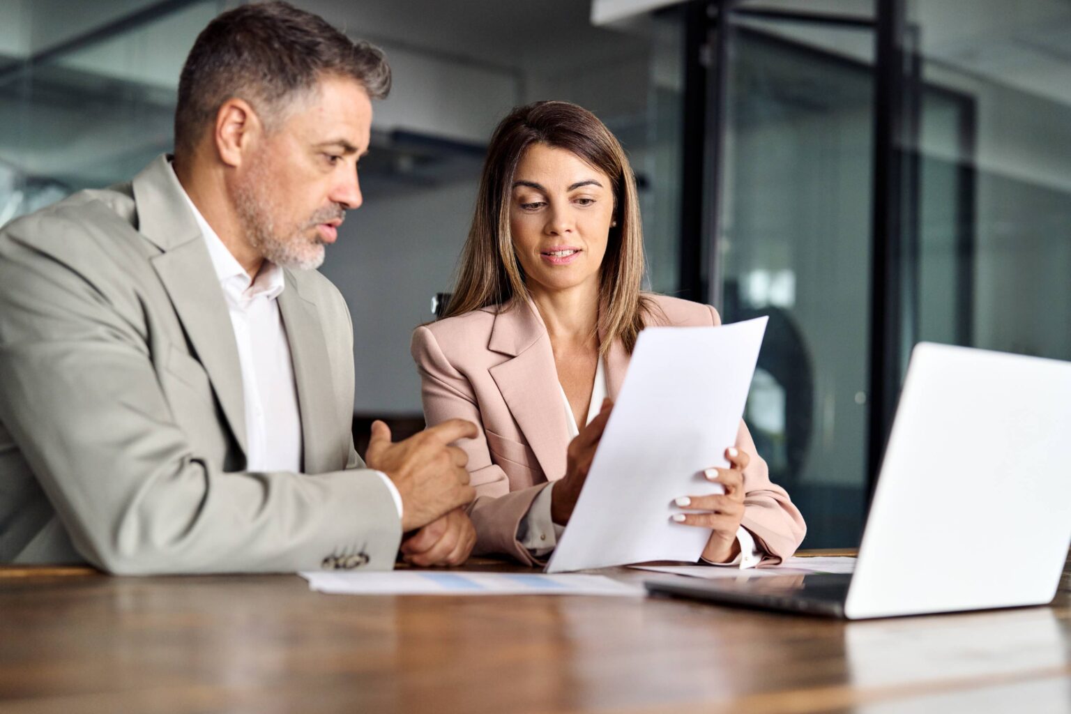 Two business professionals sit at a conference table in a modern office environment. Both are dressed in business attire—a man in a light grey suit and a woman in a blush pink blazer. The woman holds a document while the man gestures in discussion. A laptop is open on the table next to them, suggesting a meeting or collaborative work session. Their faces are intentionally blurred for privacy.