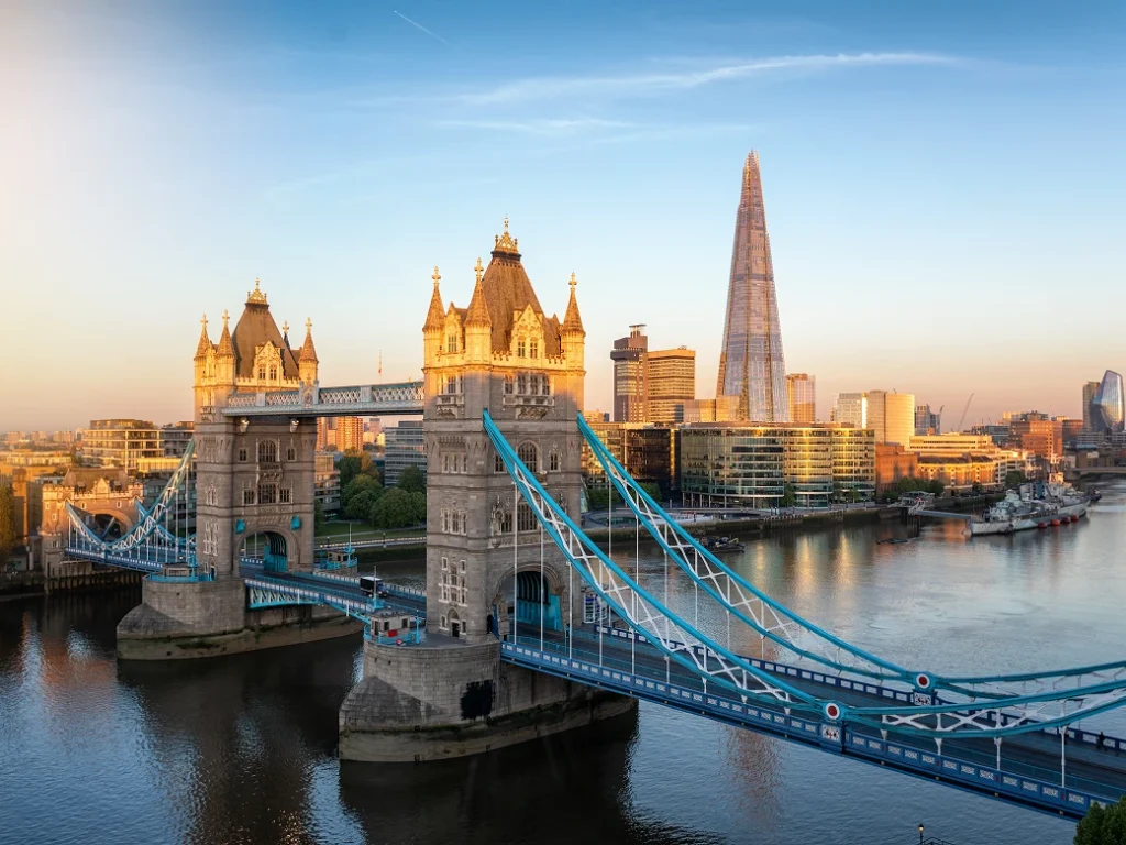 London's Tower Bridge over the River Thames at sunrise, with the Shard skyscraper and city buildings in the background.